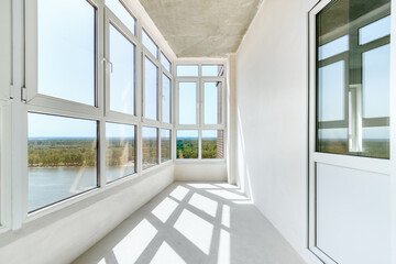 A sunny balcony with the panoramic windows in a new residential building on the high floor. A light loggia with white plastered walls without decoration - obrazy, fototapety, plakaty