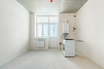 The interior of an undecorated kitchen room in a new residential building with the white plastered walls. A gas-stove and a wall mounted water heater in the empty room