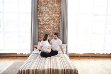Naklejka premium Portrait of happy husband and pregnant wife sitting on white bed, looking at each other at home with big window. Pregnancy wellbeing relationship support concept. Copy space, special family moments