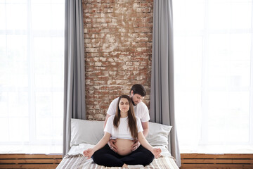 A pregnant woman sits in a lotus position with closed eyes and meditates on a bed while her husband...