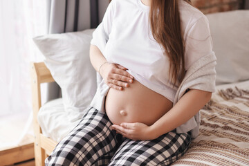 Close-up pregnant woman in home outfit: a white T-shirt and checkered pants sits on the bed, hugs her tummy. Prenatal, Baby expecting concept, studio, copy space.