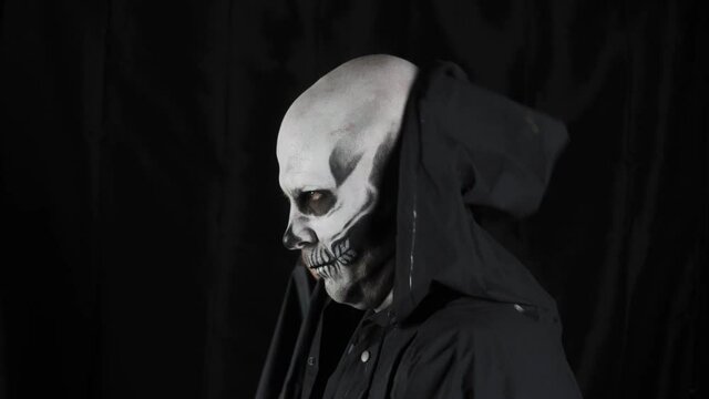 Man with make-up skeleton and black hood on a dark background. A man takes off his hood showing a skull. Halloween or horror theme.