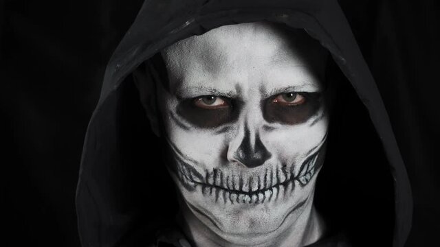 Man with make-up skeleton and black hood on a dark background. A man takes off his hood showing a skull. Halloween or horror theme.