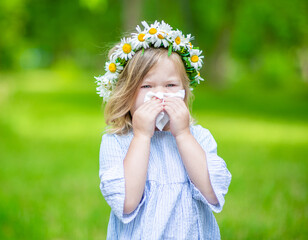 Portrait of a girl wearing wreath of daisies blowing her nose in summer park, allergic to bloom...