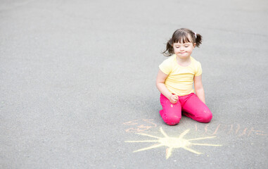 Smiling girl with syndrome down draws the sun with chalk on the asphalt. Empty space for text