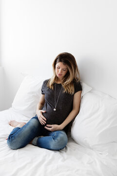 Pregnant woman sitting on white bed wearing bola necklace looking at her stomach