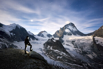 Horizontal snapshot of incredible blue sky full of stars and the Moon over cool eternal snowy peaks of Pennine Alps in Switzerland, a tourist standing and watching the beauty Dent Blanche