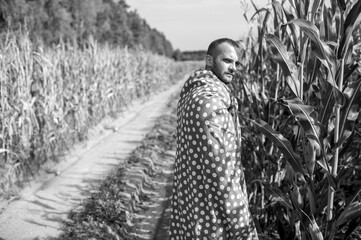 Black and white picture of beautiful caucasian man with short dark hair in wrapped in big red and white coverlet walks to the big cornfield relaxes and thinks about something