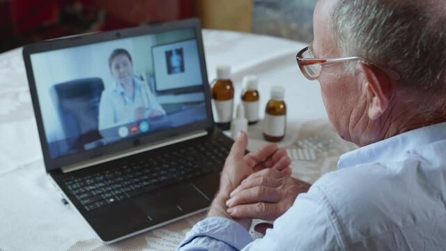 poor health, old male patient pensioner with glasses for vision consults doctor online on video call using modern technology, elderly man measures his pulse while sitting at table with laptop while