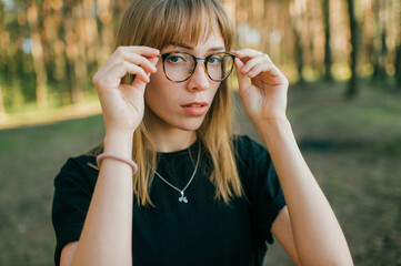 Portrait of beautiful young girl with short fair hair, blue eyes, black glasees in black jacket poses for the camera in the park