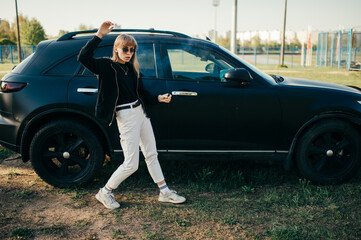 Beautiful young caucasian woman in white jeans, white trainers and black jacket stands near old black car outside