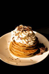 Banana pancakes with whipped cream and grated chocolate on a white plate on a black background. Vertical photo