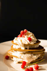 Banana pancake with red currant berries and whipped cream on a white plate, on a black background