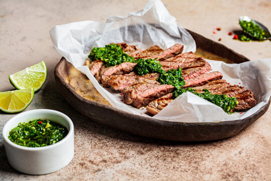 Sliced grilled beef steak with chimichurri sauce on dark dish, dark background.