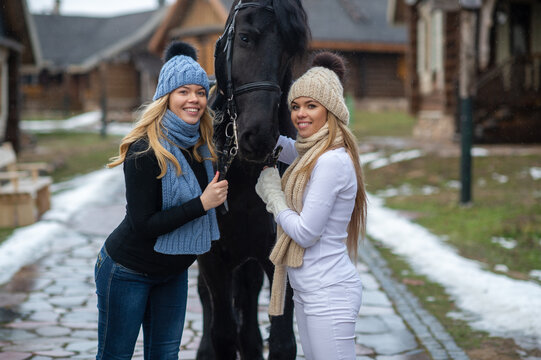 Picture of two young cheerful female riders in warm jackets, trousers, boots, scarfs and knitted hats with a horse outside