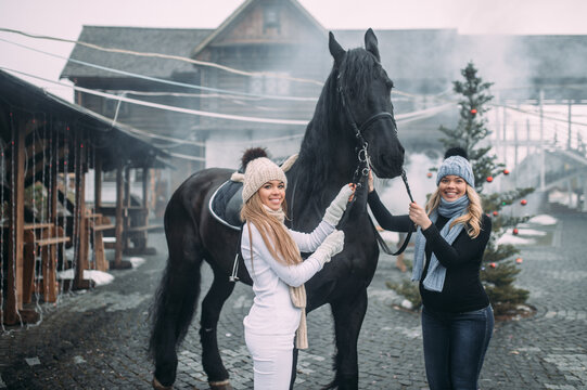 Picture of two young attractive sisters in warm jackets, trousers, boots, scarfs and knitted hats with a horse outside