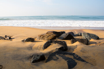 Fototapeta na wymiar natural seascape shot of a wave crushing on the smooth yellow sand beach surface with black rocks in the front and a blue sky on the horizon. Morning sunrise at Pitiwella beach, Sri Lanka
