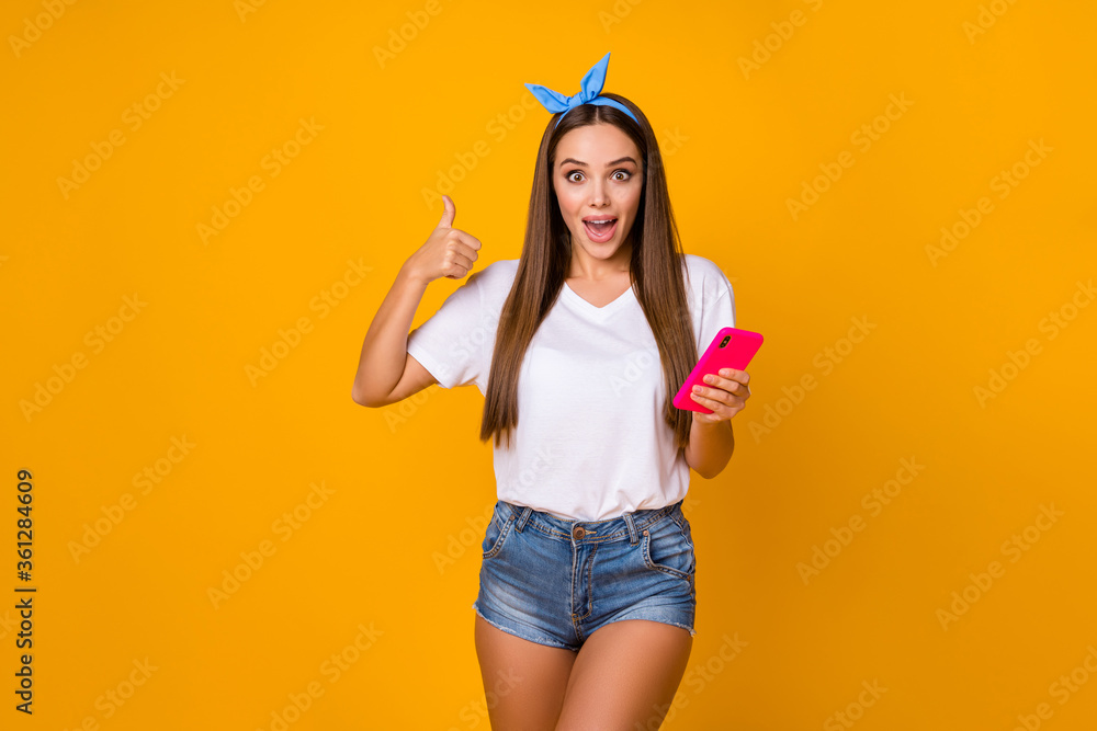 Sticker Astonished surprised girl promoter use smartphone show thumb up sign recommend incredible ads promotion impressed wear white blue headband denim jeans isolated bright shine color background