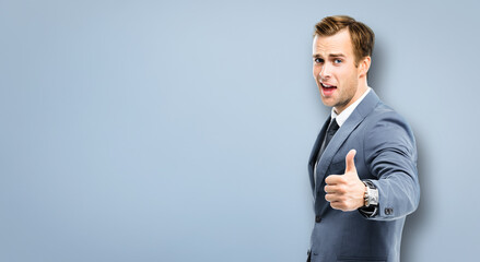 Portrait image - excited businessman showing thumbs up like hand sign gesture, in confident suit, standing over grey background. Amazed man. Copy space for some text or imaginary.