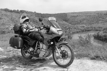 Motorcycle driver resting after a long and difficult road, lies on a motobike, wearing a turtle jacket, body armor. on top. View of the forest and river. Black and white