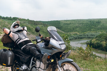 Close view of a motorcycle driver resting after a long and difficult road, lies on a motobike, wearing a turtle jacket, body armor. on top. View of the forest and river. The concept of freedom