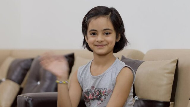 Adorable Indian kid/child doing a hello gesture while looking towards the camera. A cute little girl in casual summer wear smiling and waving her hand while sitting on a couch - expressions and ges...
