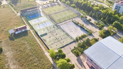 Aerial view of tennis court. Audience stand, football field on the side and other tennis field can be seen. Tennis players are training.