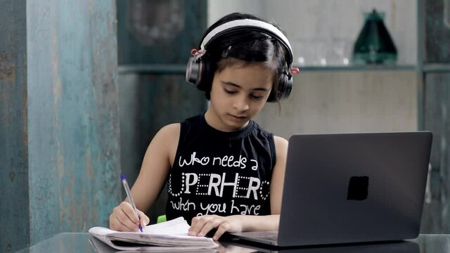 A cute little girl in black headphones taking online school classes on her laptop. Indian kid sincerely attending school lectures online due to the outbreak of coronavirus - distance learning concept