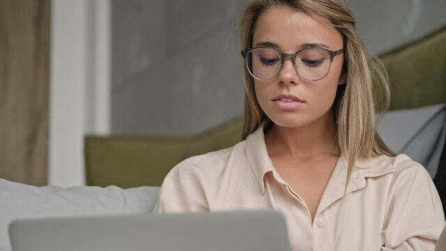 Concentrated attractive blonde woman in eyeglasses using laptop computer while sitting on the floor at home