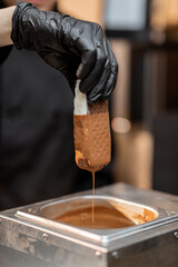 Chef in black workwear dips ice cream into the hot melted chocolate, making ice cream on a stick with toppings