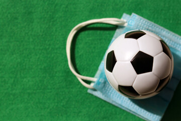Top view of football or soccer ball with blurred medical face mask on green background. The concept of new normal for sporting event and football competition during Covid-19 or Coronavirus. 