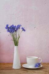Cornflowers and coffe cup on a pink wall on a brown wooden table. Cornflowers in a vase. Ceramic tableware. White vase. Classic. Relax