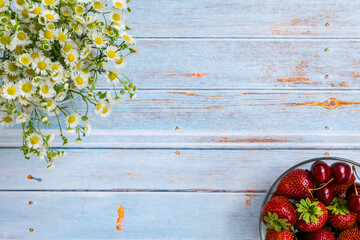 Composition. Strawberries, cherries in a glass plate. Chamomiles. Strawberries, cherries and daisies on a blue wooden background.