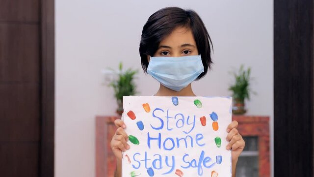 Portrait of a young girl wearing a surgical mask during the Covid-19 pandemic time. Cute Indian kid showing a drawing with the inscription of "Stay Home  Stay Safe" - campaign for coronavirus preve...