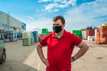 A man in a black mask and a red shirt is standing at a construction warehouse outside with his hands on his hips.