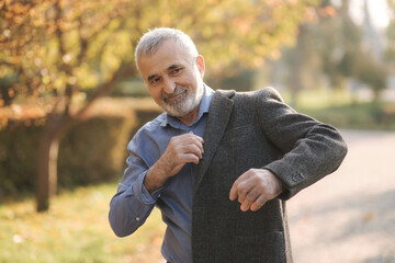 Handsome elderly man putting on a gray jacket. Old gray-haired bearded man walk in the autumn park. Yellow background