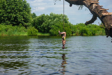 The boy hangs on a rope with his head down towards the river.