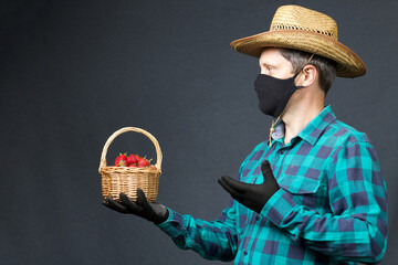 A man holds in one hand a basket with strawberries, with the other hand points to it. Farmer with a protective mask on his face. There is a straw hat on his head. Shot on a gray background.