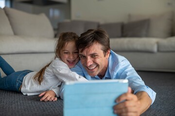 A happy smiling father and daughter enjoying time together and using a tablet for family entertainment while lying on a floor in living room at home.