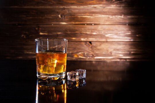 Glass of whiskey with ice cubes on black table against wooden background