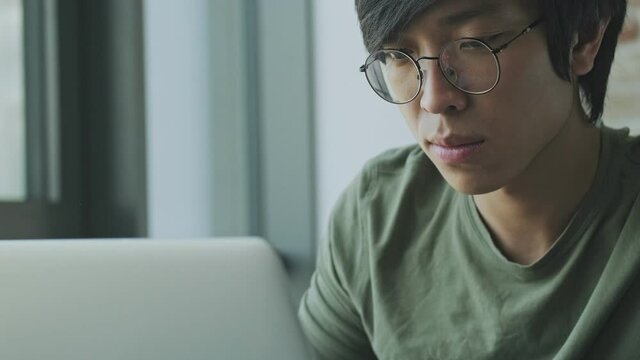 A close-up view of a calm young asian freelancer wearing eyeglasses is using his laptop computer while sitting at the table at home