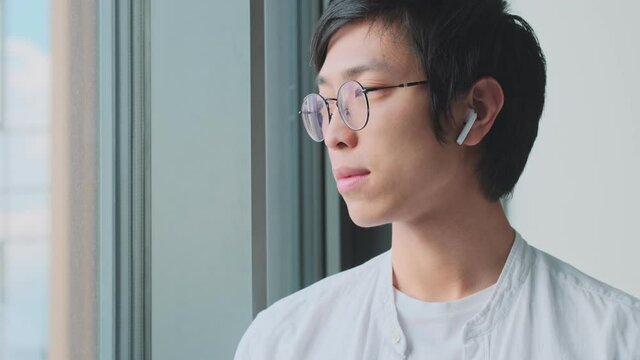An attractive young asian man with earbuds is looking out the window at home