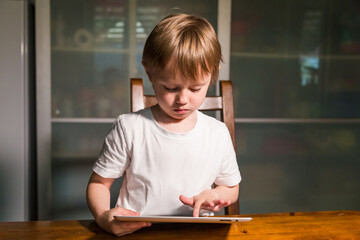 Young girl sitting at the table in the kitchen and watching cartoons on tablet. 