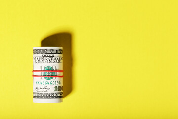 A roll of hundred-dollar American bills is tied with a red elastic band on a yellow background.