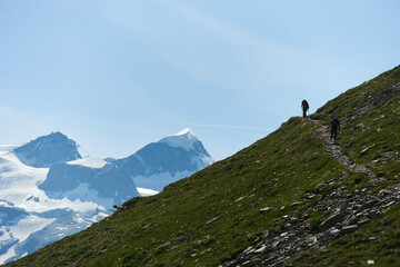 Fototapeta na wymiar Back view of two mountaineers with backpacks walking on trail using trekking sticks, beautiful mountains scenery on background. Mountain hiking, men reaching peak in summer day. Sport tourism in Alps.