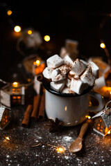 Winter hot drink: hot chocolate with marshmallows, cinnamon and anise. Christmas lights in the shape of houses as decor. Decorative snow, dark wooden background. Festive mood, cozy home atmosphere