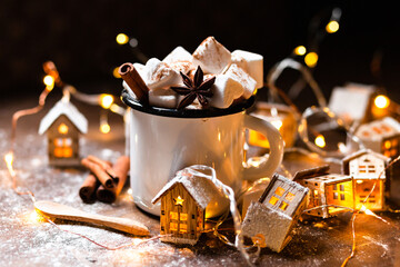 Winter hot drink: hot chocolate with marshmallows, cinnamon and anise. Christmas lights in the shape of houses as decor. Decorative snow, dark wooden background. Festive mood, cozy home atmosphere