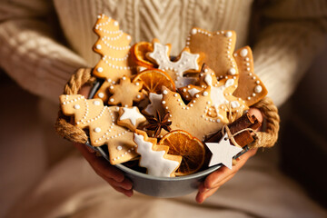 Young girl in knitted white sweater is holding a basket with homemade gingerbread cookies decorated with snow white icing. Anise and cinnamon as decor. Close up. Christmas lights on background