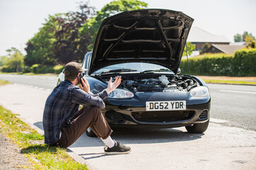 Young man next to his car, depressed and angry on the mobile phone telephone call after the car has broken down on the roadside waiting for assistance to arrive and recover or fix the vehicle