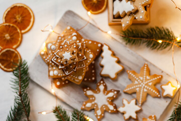 Delicious homemade gingerbread cookies with white icing. Christmas lights, branch of fir tree, dry orange slices. White wooden background, flat lay, top view. Festive holiday atmosphere, family time